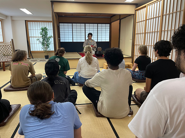 View of students sitting on floor cross legged from back of the room with tea ceremony hostess facing them