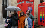 Five students on a street in London standing in front of a bright red old fashioned callbox. The students are wearing rain gear and one of them is holding an umbrella up