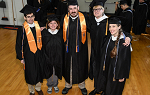 Five students in caps and gowns stand with their arms around each other and look up at the camera