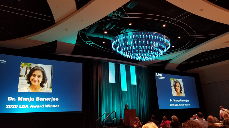 A long distance shot of Manju Banerjee speaking the podium in a ballroom at the LDA Conference in Orlando after accepting her award. Two video screens on each side her show her picture with text that reads 